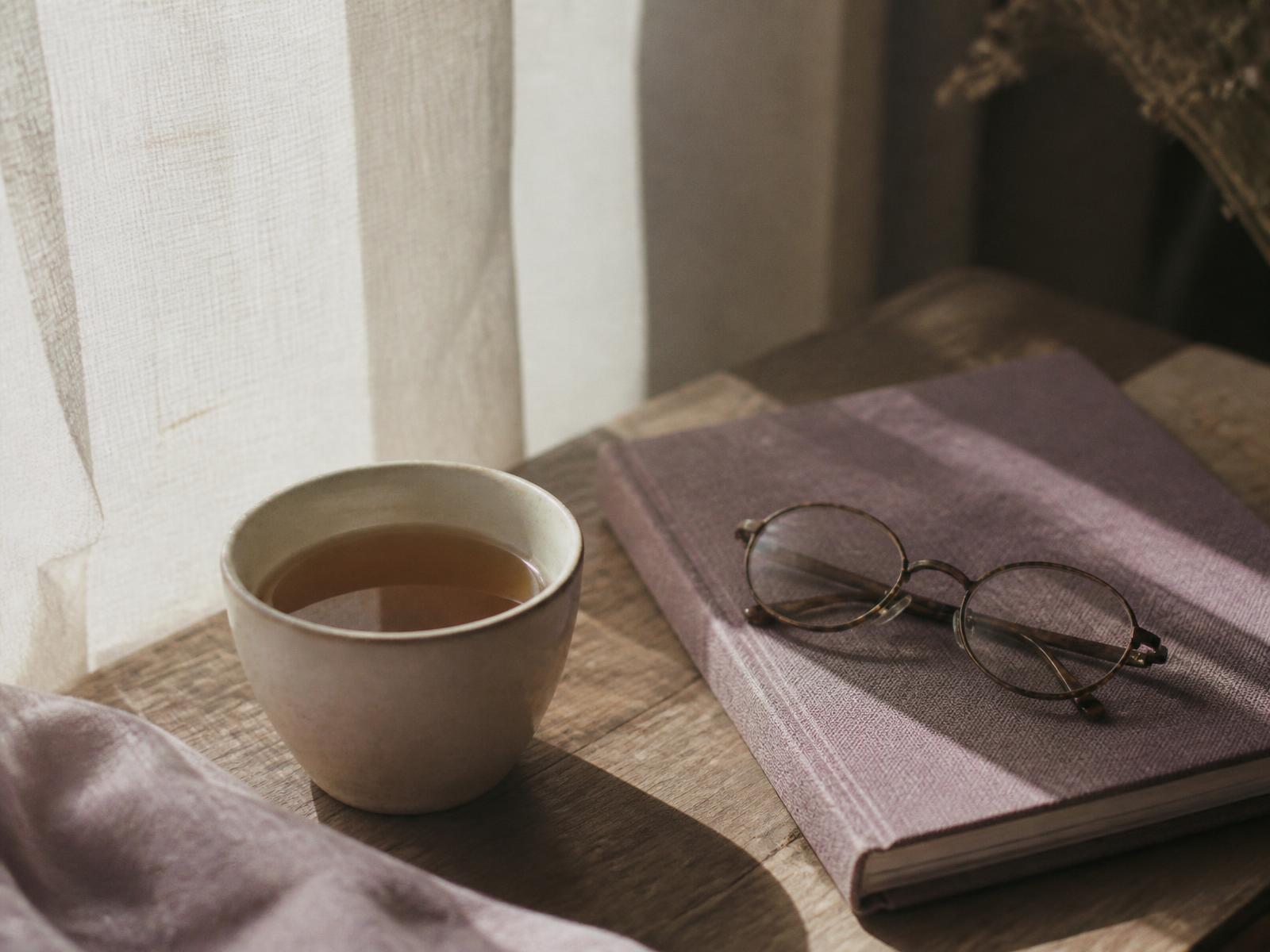 Mesa de madera con una taza de té, un cuaderno y unas gafas junto a una ventana con luz suave